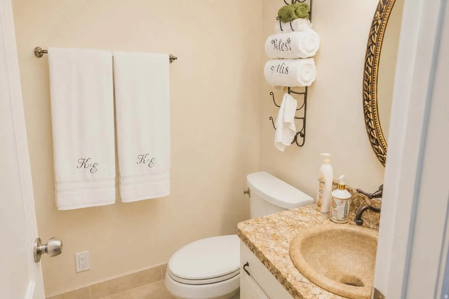 Powder room featuring a stone countertop vanity with a bronze-finish faucet, an oval mirror with an ornate frame, ceramic tile flooring, and a towel bar