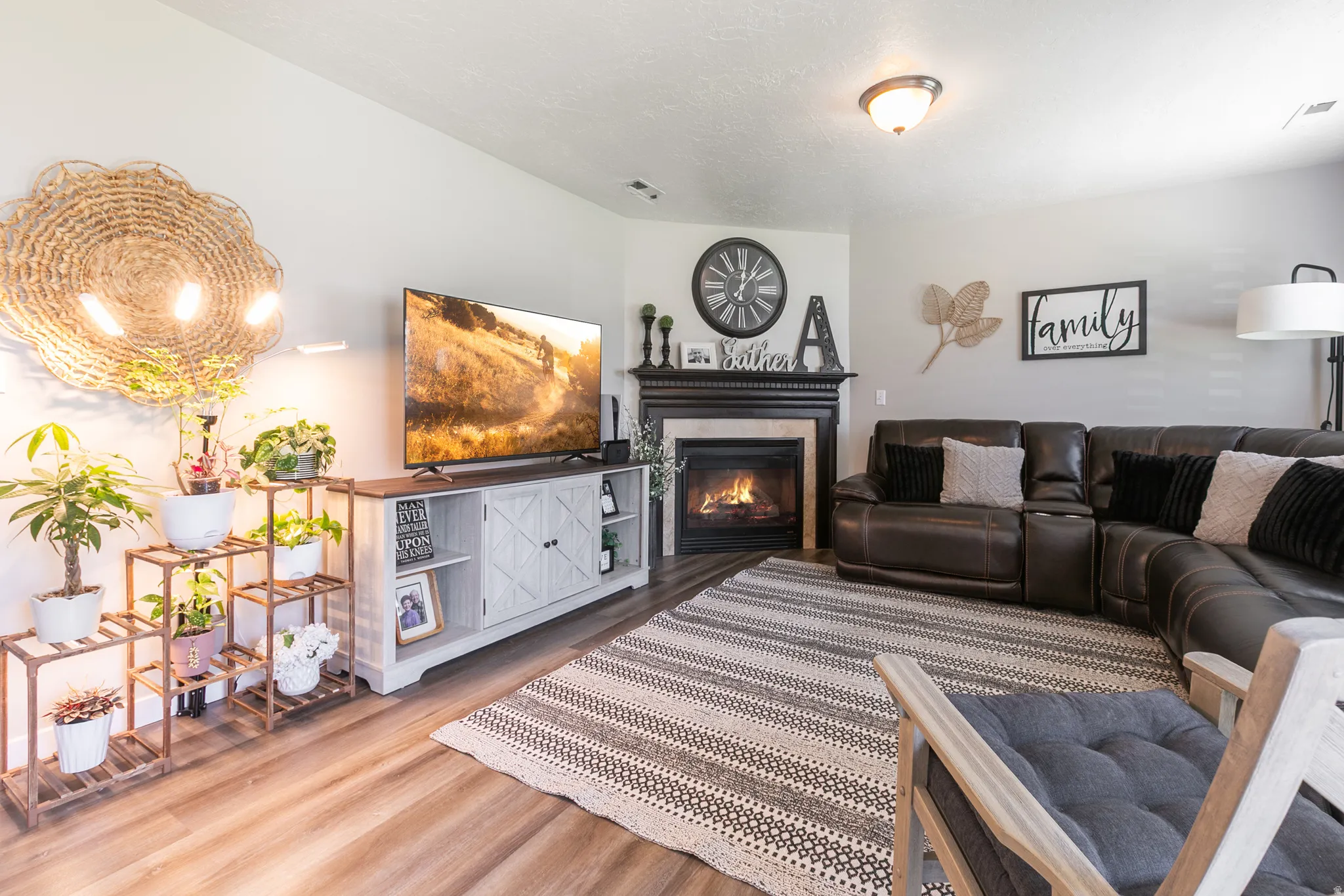 Living area with wood-finish flooring and a corner fireplace featuring a dark wood mantel and light-toned surround