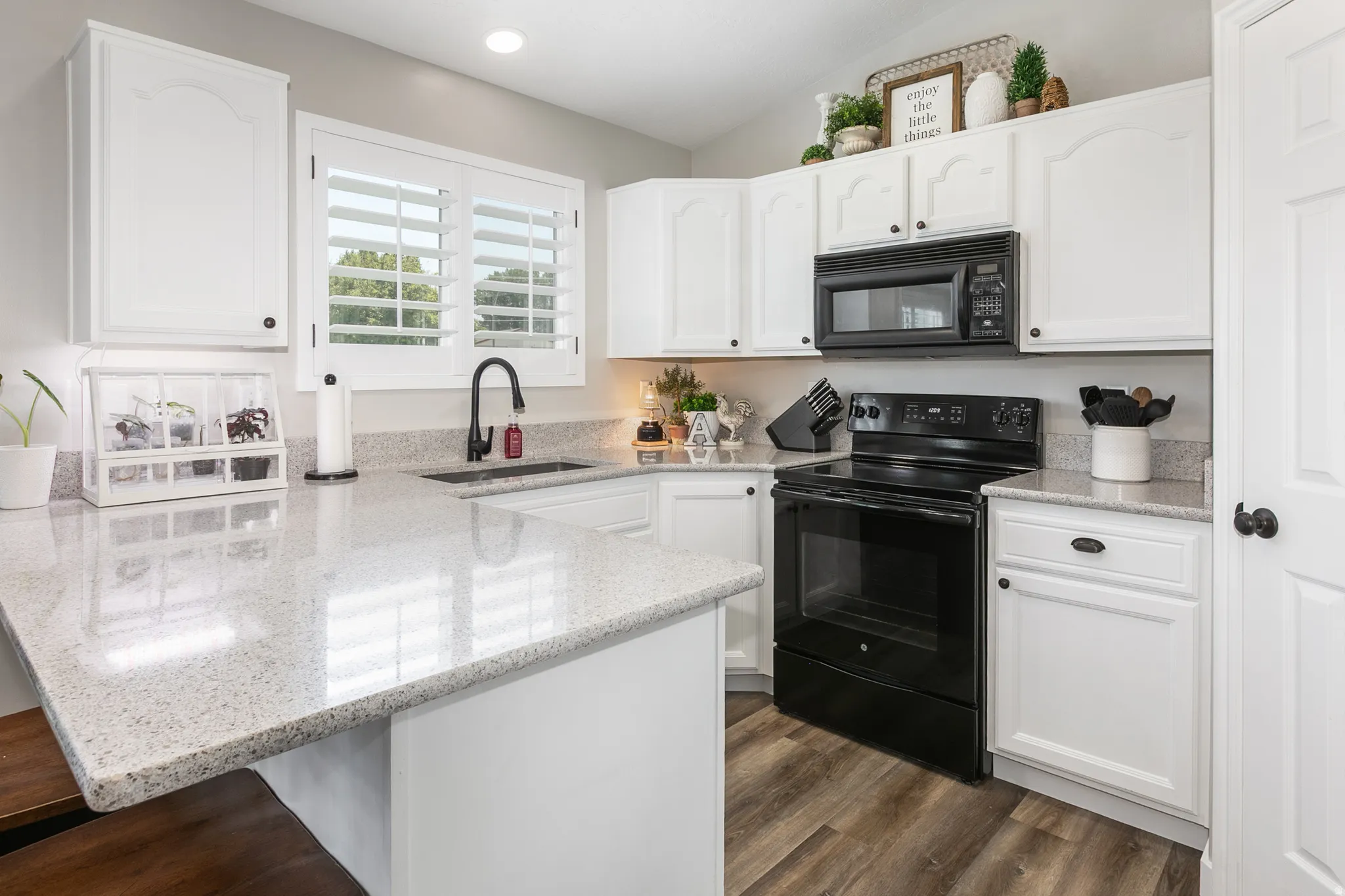Modern kitchen featuring white cabinetry, speckled countertops, stainless steel sink with a black faucet, black appliances, and wood-finish flooring