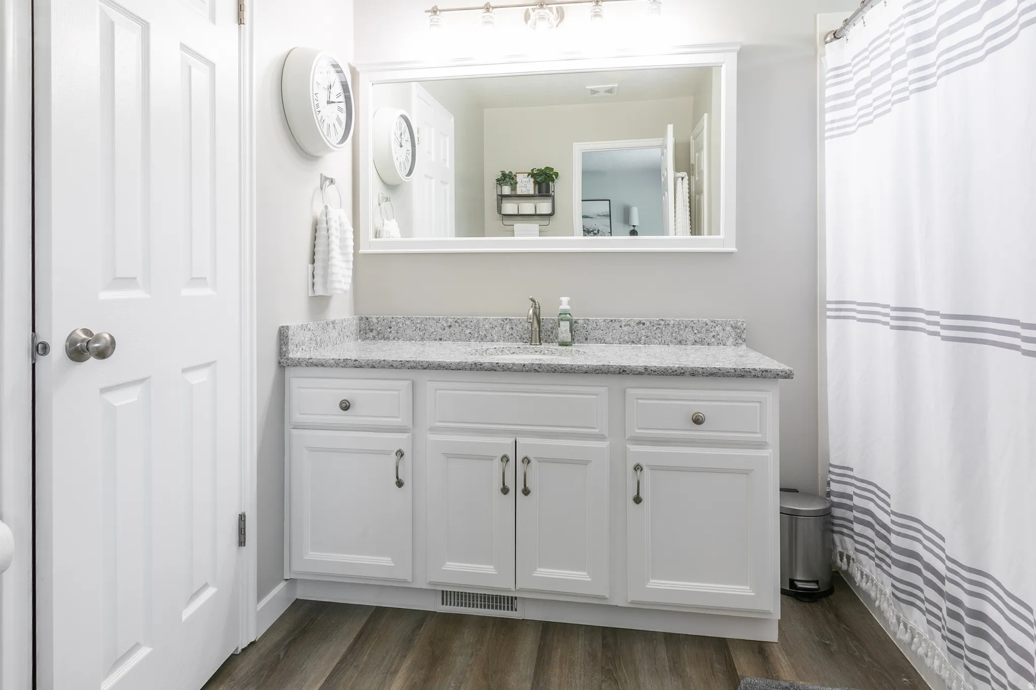 Bathroom featuring a white vanity with ample storage, a speckled countertop, a large framed mirror, and wood-finish flooring