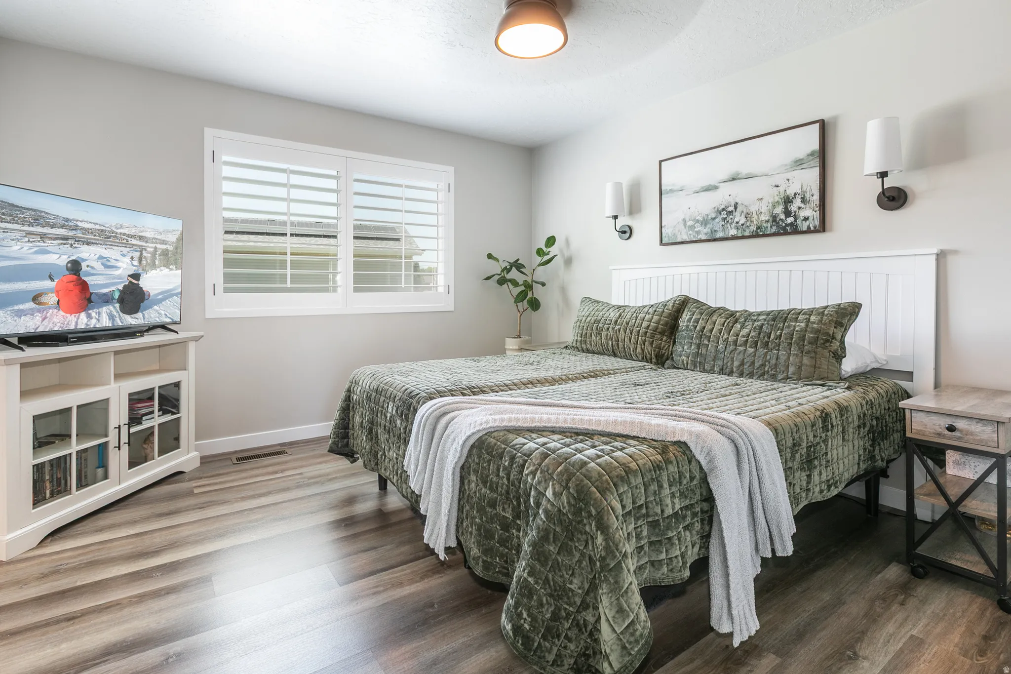 Bedroom featuring wood-finish flooring, white plantation shutters, a white paneled headboard, and dual wall sconces