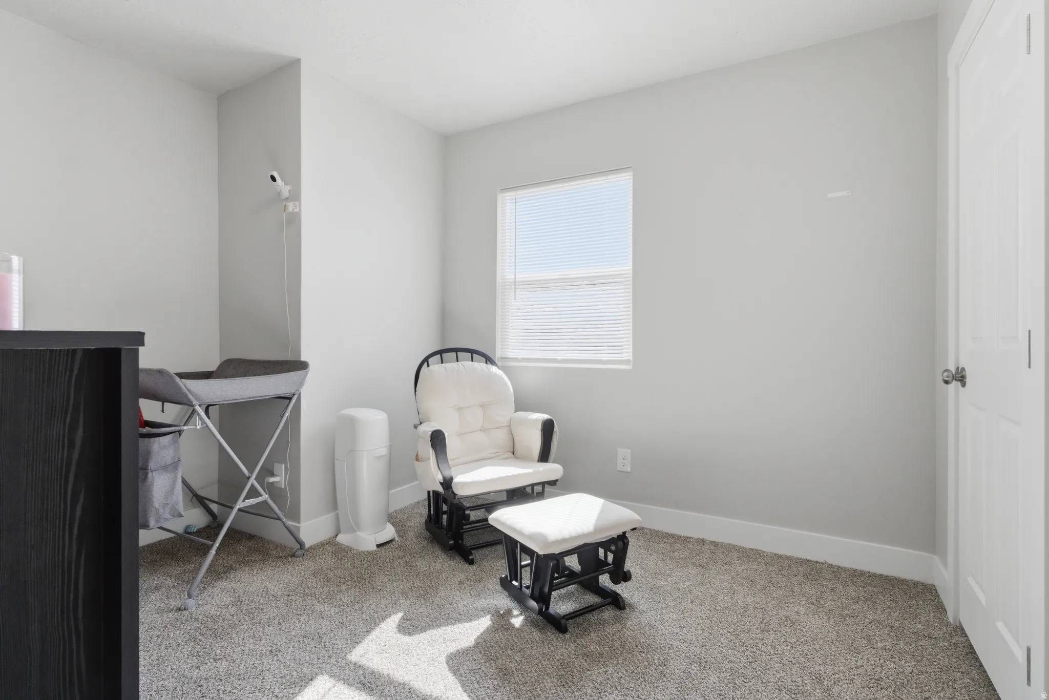 Carpeted room with light gray walls, a single window featuring horizontal blinds, and a white paneled door with a brass-finish knob