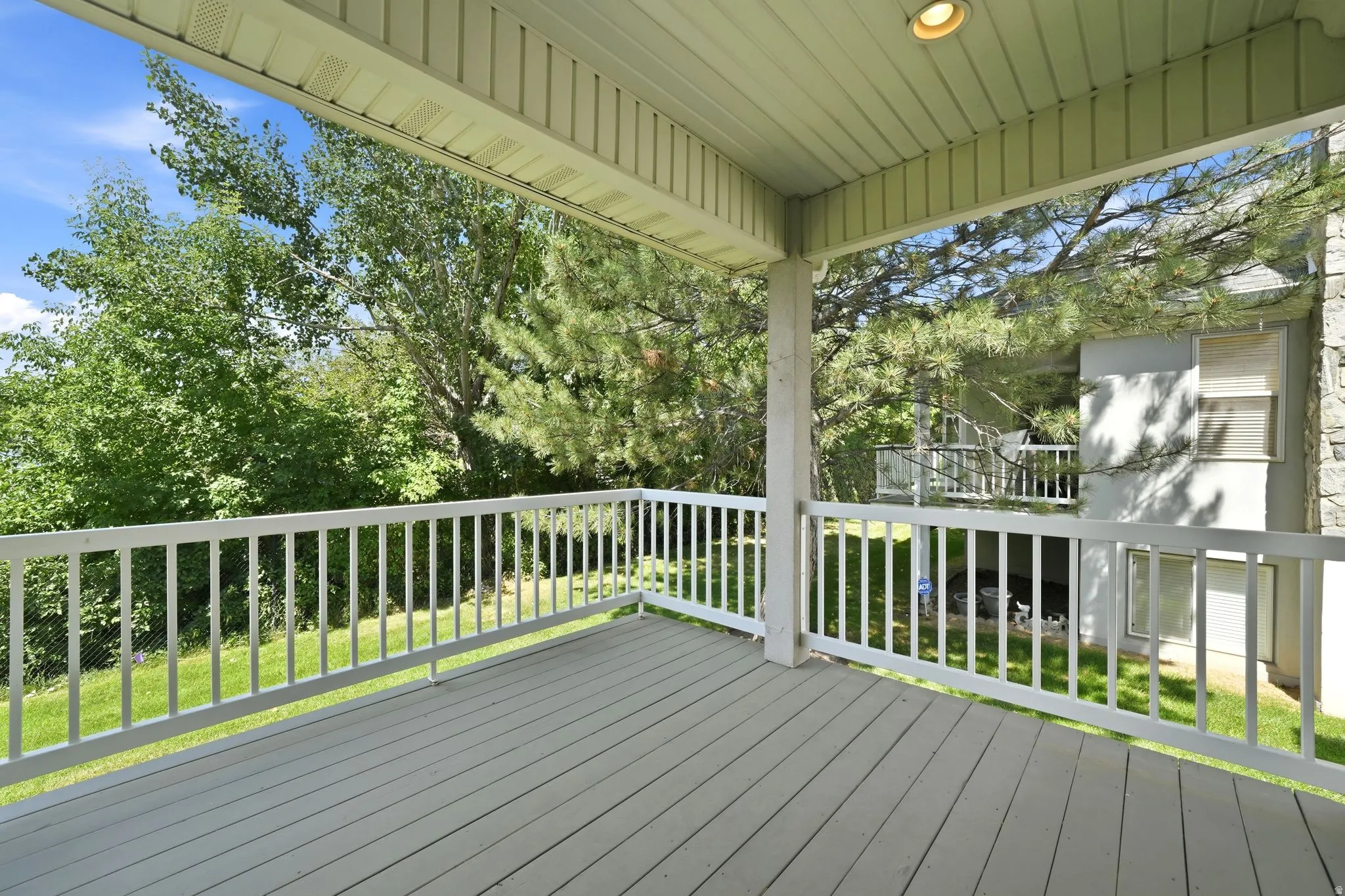 Covered outdoor deck with light gray wood-finish flooring, white railing, and a textured overhead ceiling featuring a recessed light fixture