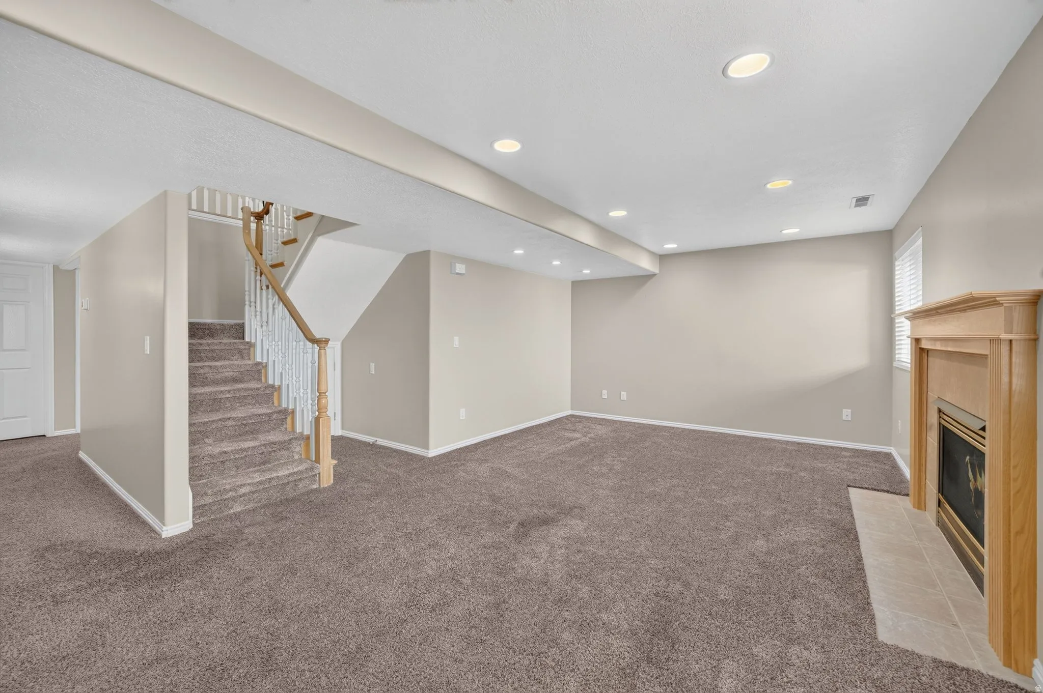 Spacious room featuring neutral carpeting, recessed lighting, a wood-surround fireplace with a tiled hearth, and a carpeted staircase with a wood handrail and white balusters