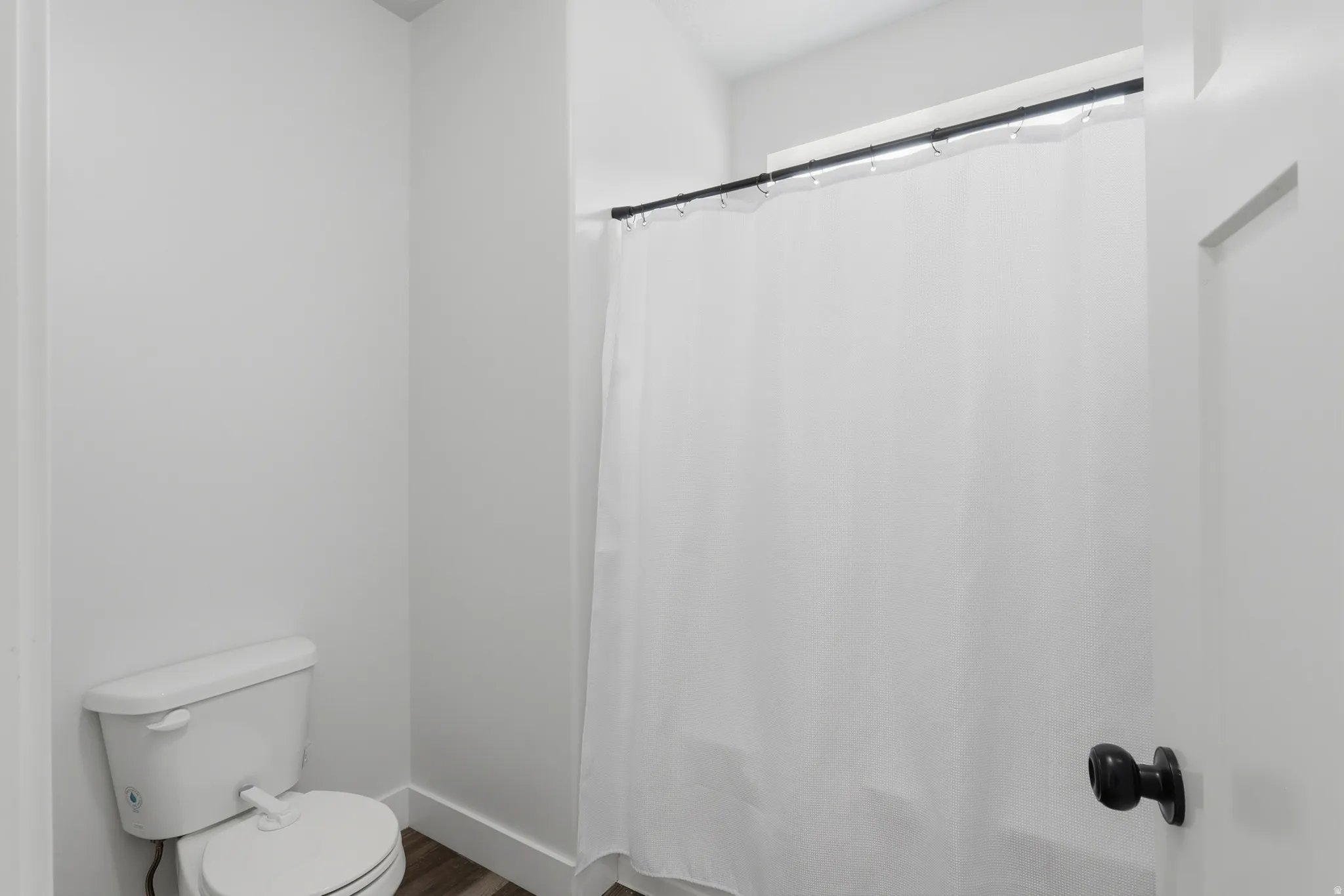 Bathroom featuring a white toilet, wood-finish flooring, and a shower with a black curtain rod