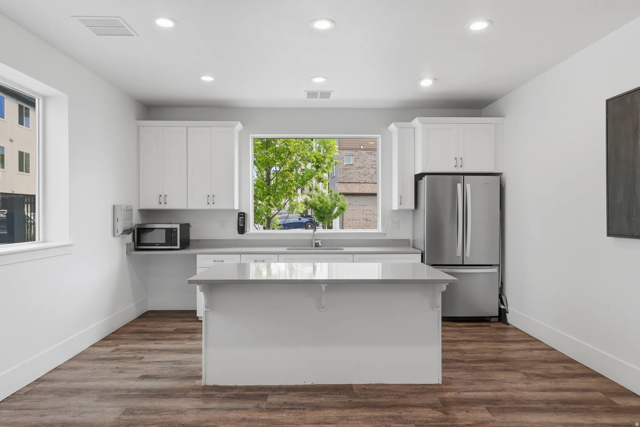 Modern kitchen featuring white cabinetry, light-toned countertops, a central island, stainless steel refrigerator, and wood-finish flooring