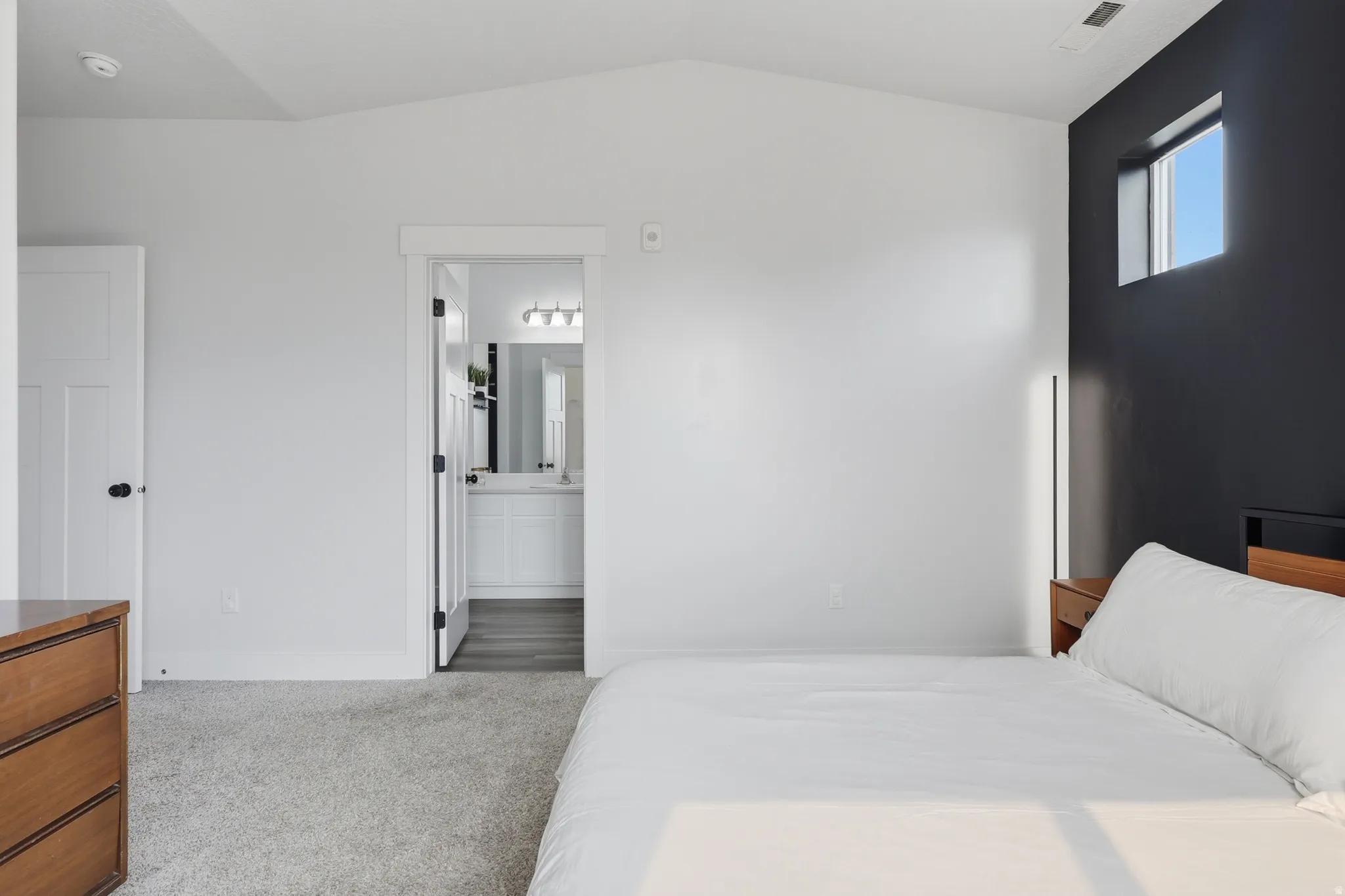 Vaulted ceiling bedroom featuring an accent wall with a high window, soft gray carpeting, and an adjoining bathroom with a vanity and mirror