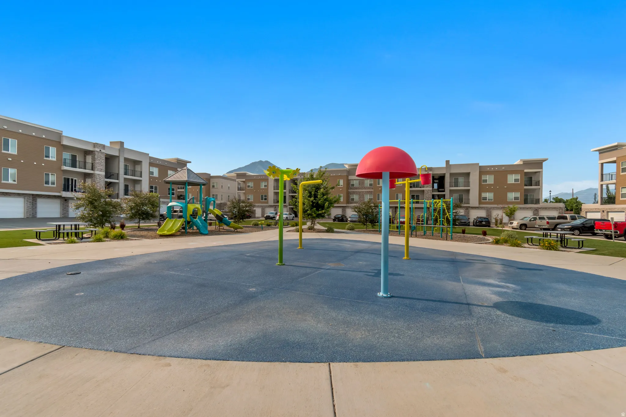 Community splash pad featuring colorful play structures, an adjacent playground, green space, and communal picnic tables, all surrounded by multi-story apartment buildings with balconies
