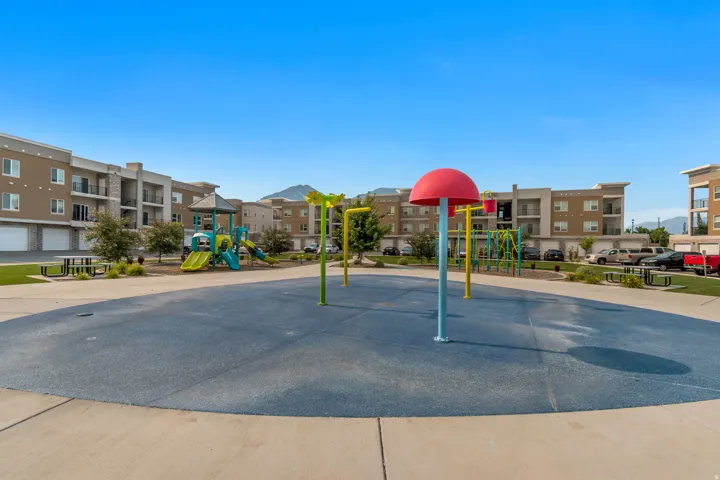 Community splash pad featuring colorful play structures, an adjacent playground, green space, and communal picnic tables, all surrounded by multi-story apartment buildings with balconies