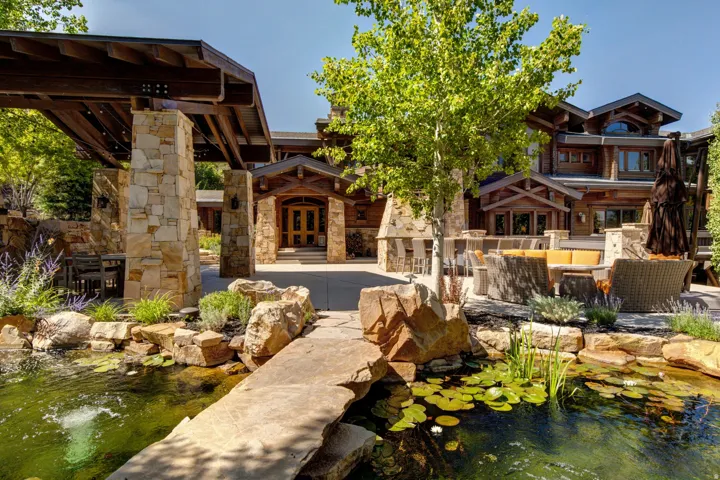 Expansive custom residence featuring a stone-clad exterior with wood accents, a stone patio with a built-in fireplace, a covered outdoor dining area, and a tranquil pond with a stone pathway