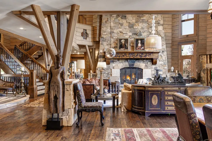 Great room featuring a floor-to-ceiling stone fireplace with a carved wood mantel, exposed timber beams, wood-finish flooring, and a grand staircase with dark iron balusters