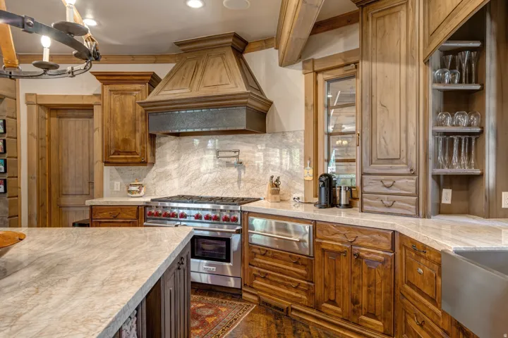 Gourmet kitchen featuring a built-in stainless steel range with a custom wood hood, light-toned stone countertops, a full stone backsplash, and wood cabinetry throughout