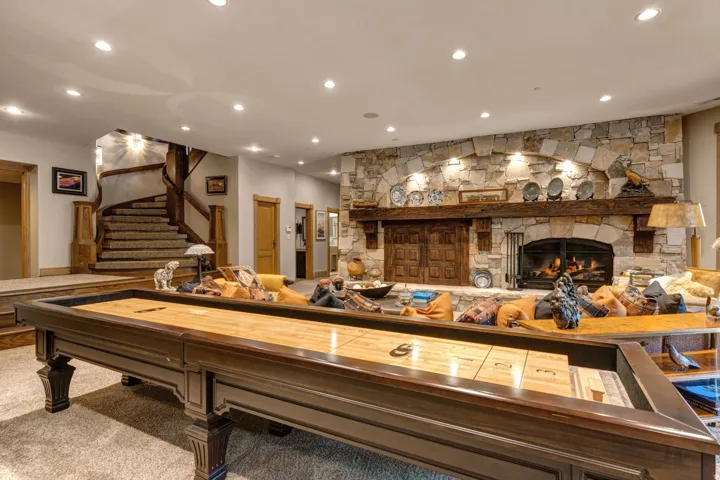 Spacious entertainment area featuring a stone fireplace with a substantial wood mantel, recessed lighting, a built-in shuffleboard table, and a carved wood spiral staircase