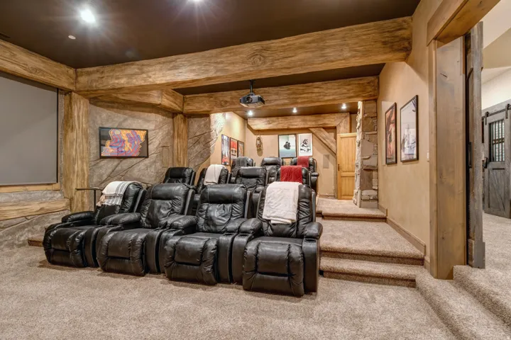 Dedicated media room featuring tiered seating, a large projection screen, rough-hewn timber beams, stone-finish walls, and plush carpeting