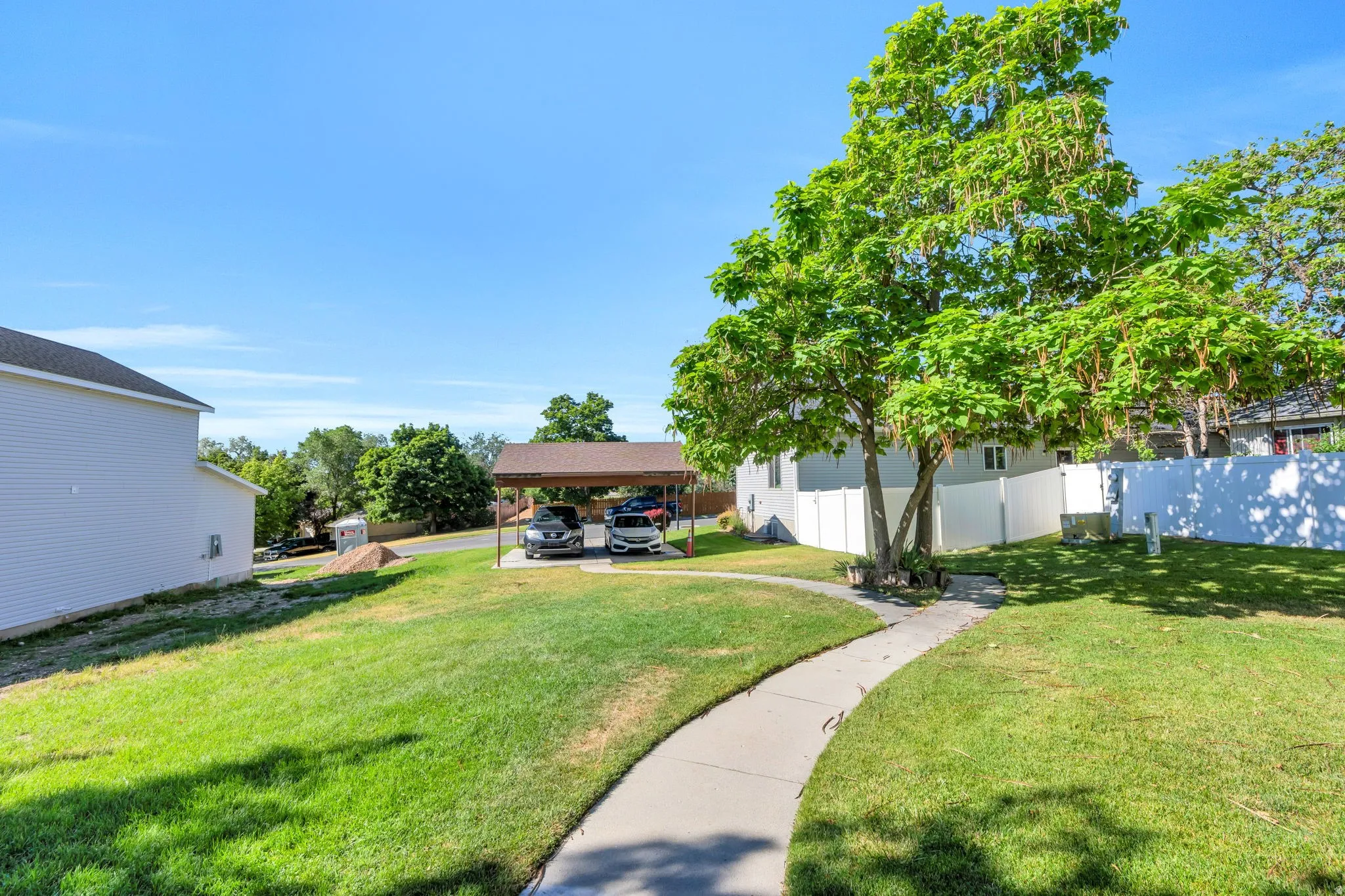 Spacious lawn featuring a meandering concrete pathway, mature tree, white privacy fencing, and a two-car carport