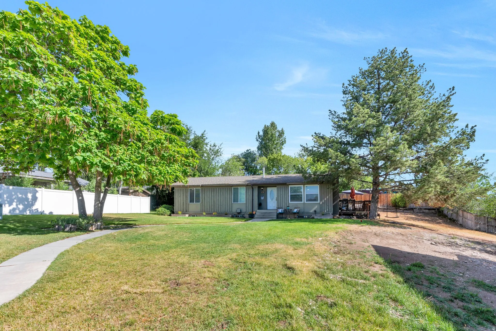 Ranch-style residence featuring a prominent mature tree, a curved concrete pathway leading to the entrance, an expansive lawn area, and a white privacy fence