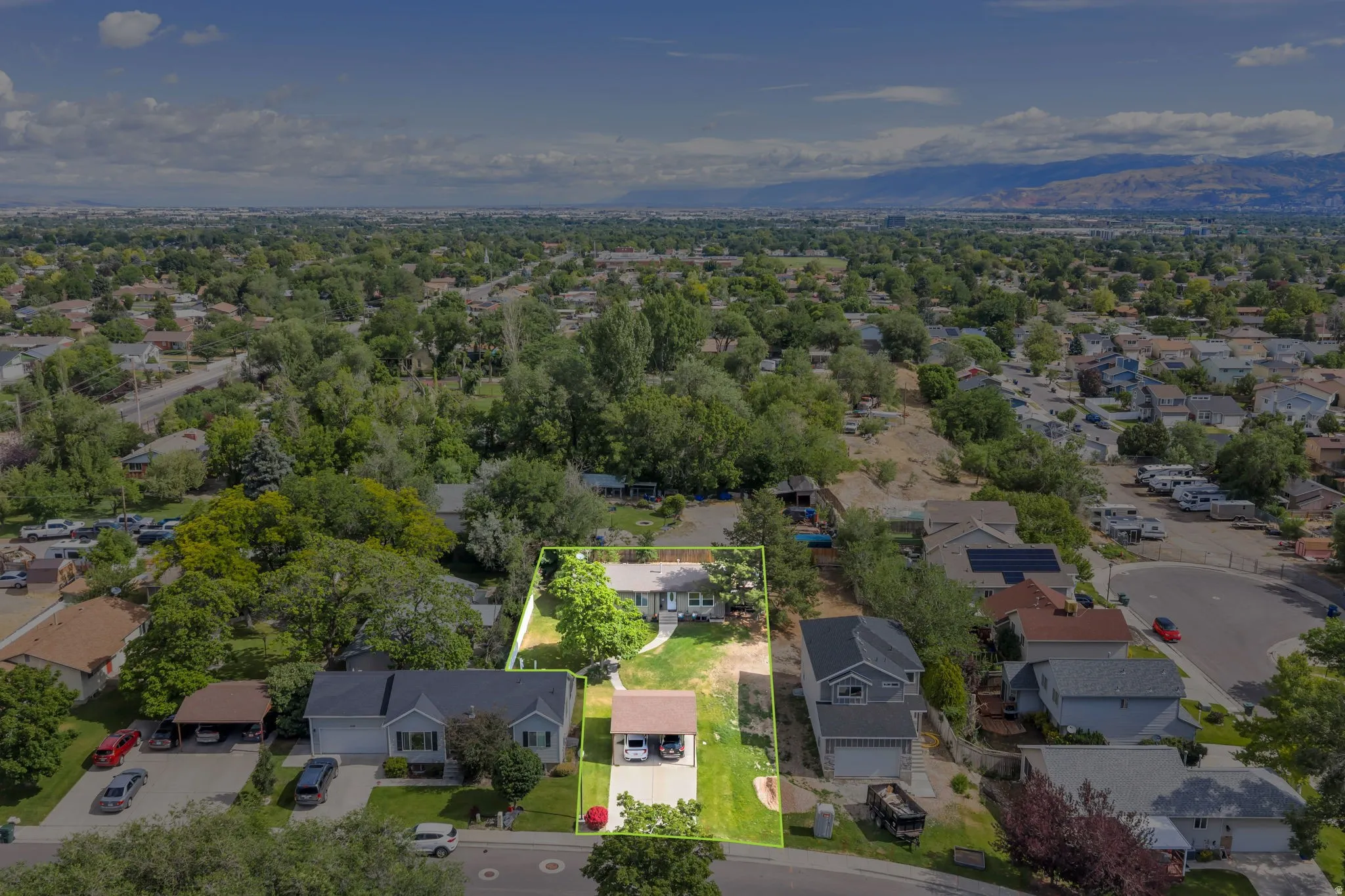 Aerial overview of a residential property featuring a light-colored exterior, lush green lawn, mature trees, and a paved driveway with a covered parking area