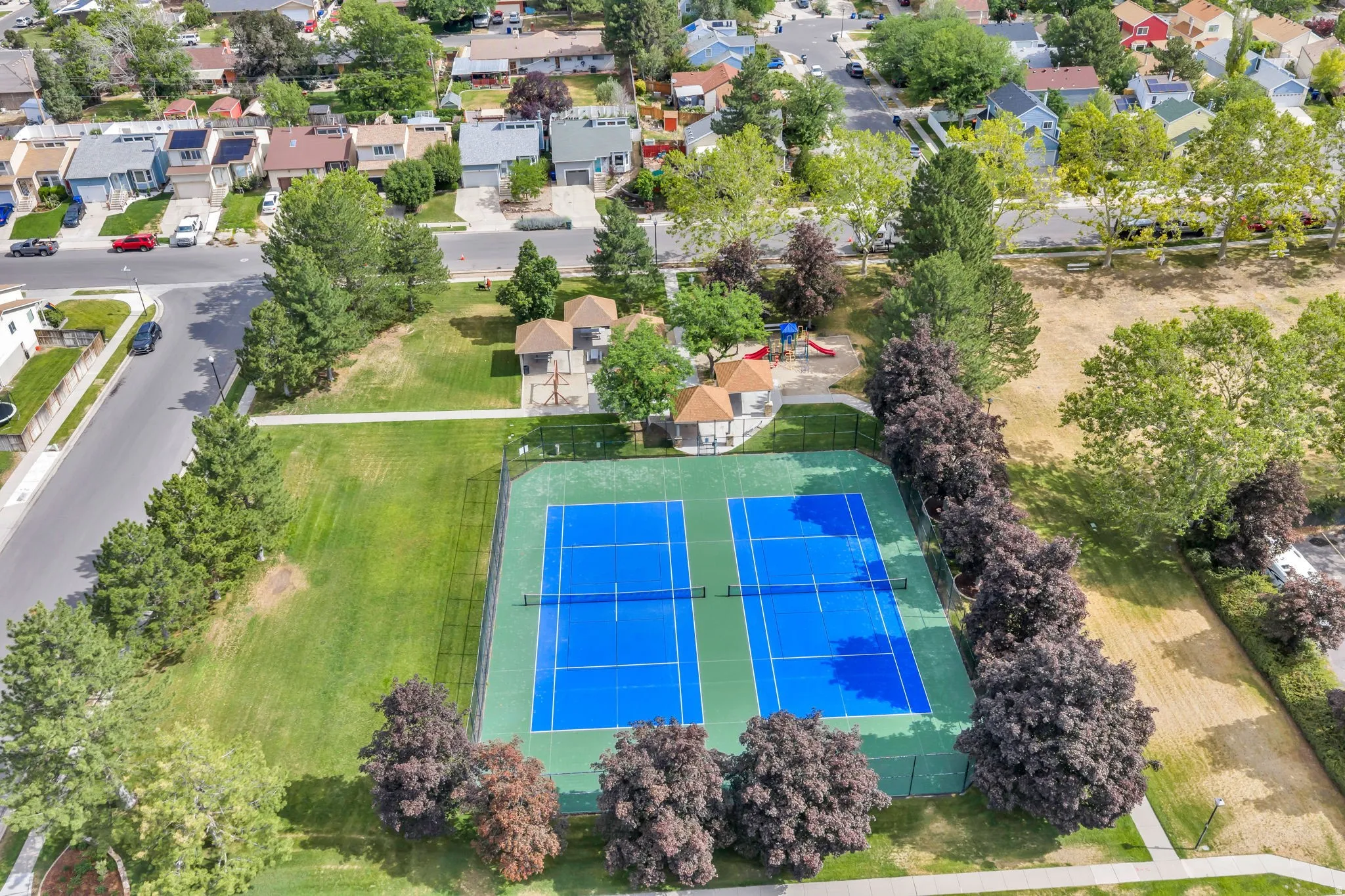 Aerial view of a community park featuring twin tennis courts, shaded picnic areas, and a children's play structure