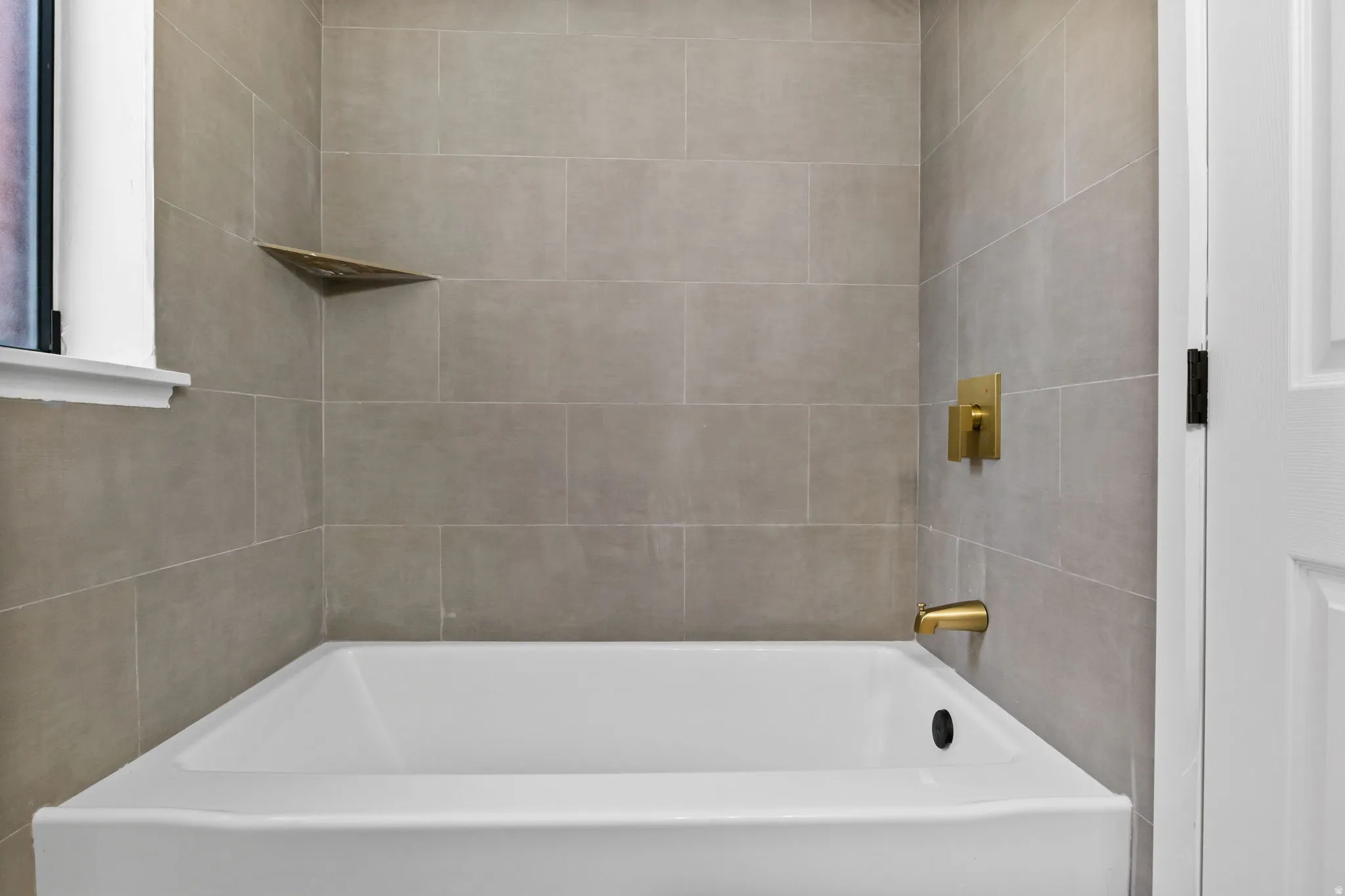 White bathtub with gray tile surround, gold-tone fixtures, and a built-in corner shelf