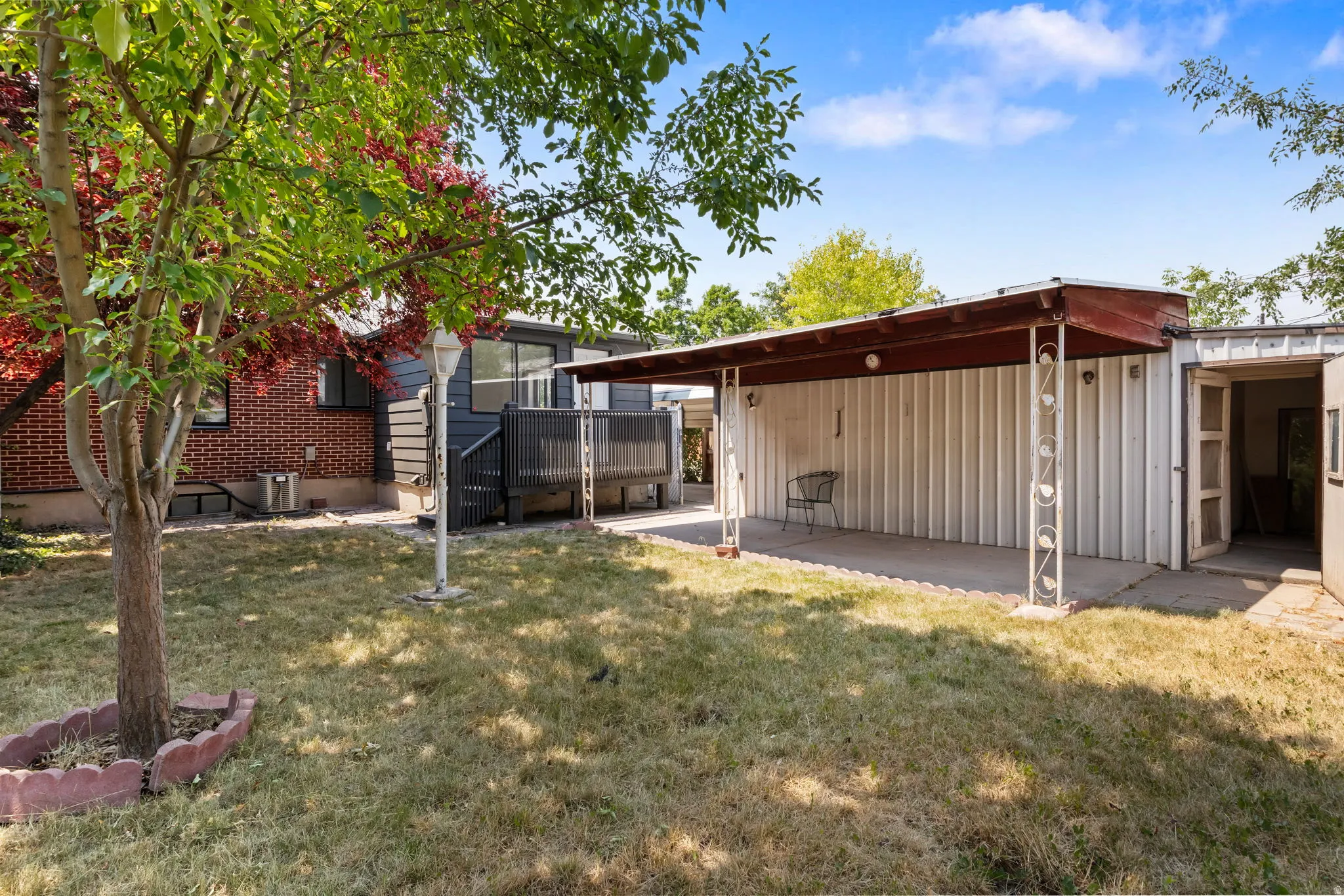 Rear exterior featuring a covered patio with decorative metal supports, an adjacent outbuilding with an open doorway, a dark-stained wood deck, and a partially visible brick exterior with horizontal siding