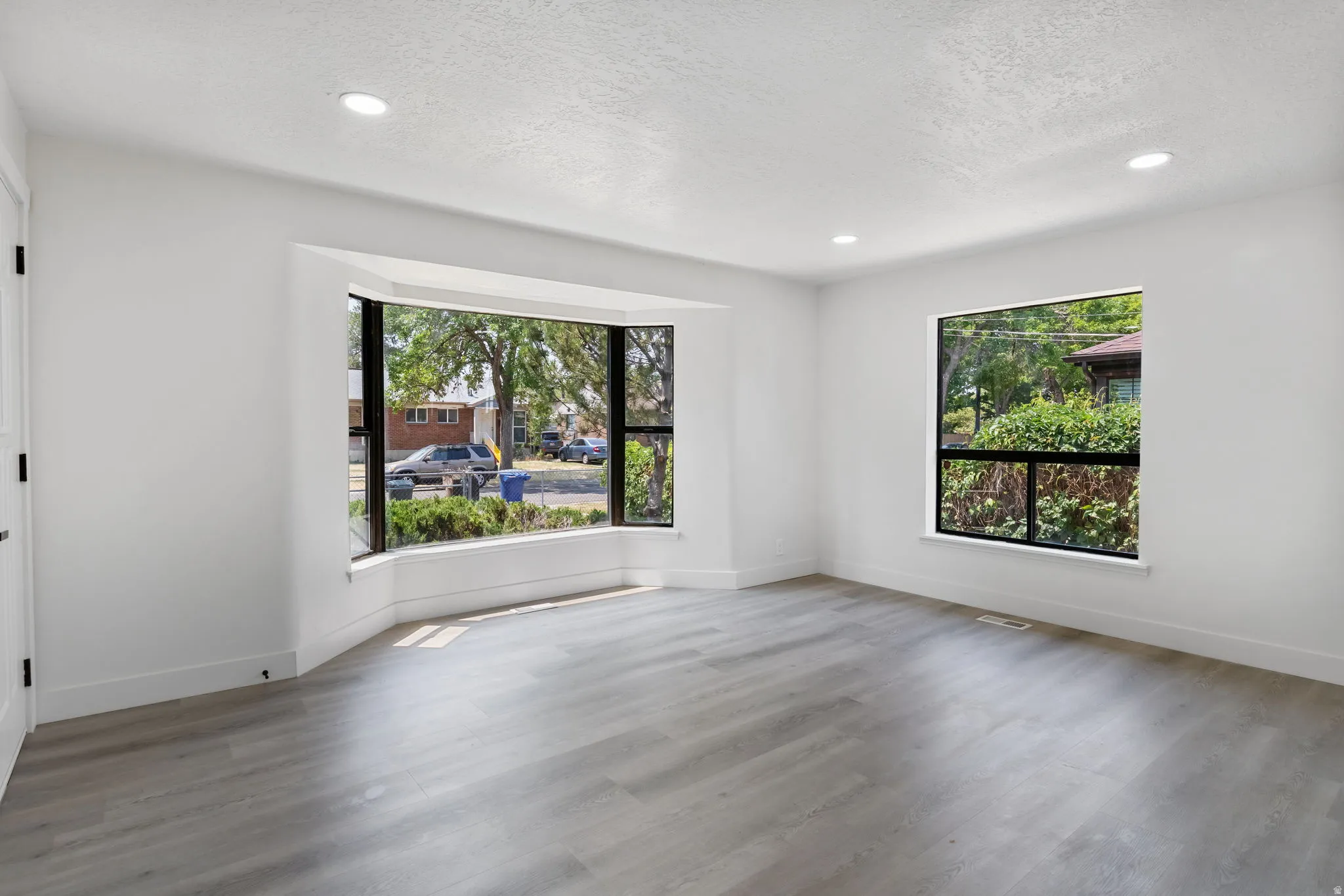 Spacious room featuring a prominent bay window with black frames, wood-finish flooring, recessed lighting, and crisp white walls