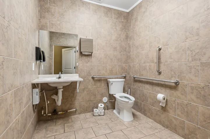Neutral-toned tiled restroom featuring a wall-mounted sink with a chrome faucet, a white toilet, and multiple stainless steel grab bars
