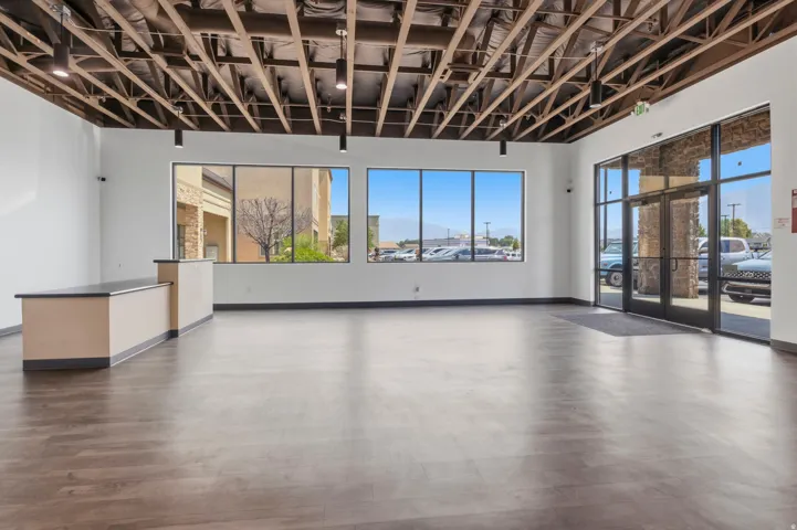 Spacious open-concept commercial interior featuring wood-finish flooring, exposed ceiling trusses with recessed lighting, expansive storefront windows, a built-in reception desk, and a glass-paneled entry door