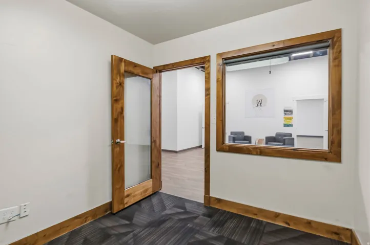 Office space featuring a wood-framed glass door, a large interior window, contemporary patterned carpet, white walls, and wood-finish baseboards