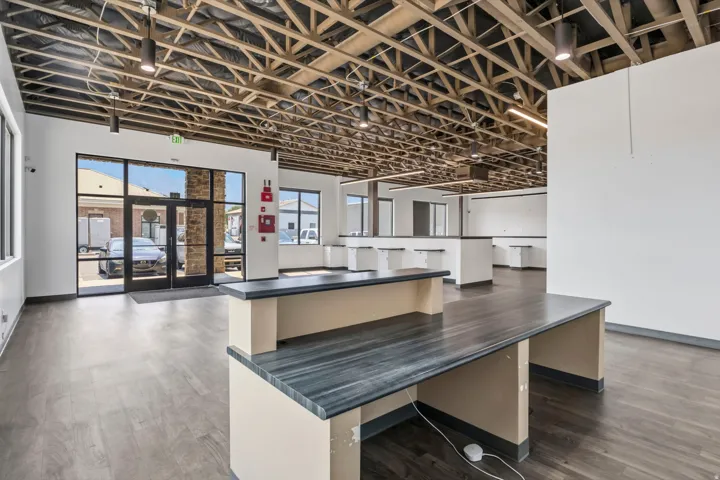 Spacious commercial interior featuring exposed ceiling trusses, recessed lighting, expansive glass entry doors with transom windows, wood-finish flooring, and built-in counters with dark countertops