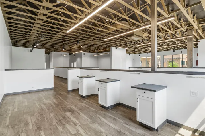 Open-concept commercial interior featuring exposed beam ceiling, linear and track lighting, wood-finish flooring, white cabinetry with dark countertops, and large exterior windows
