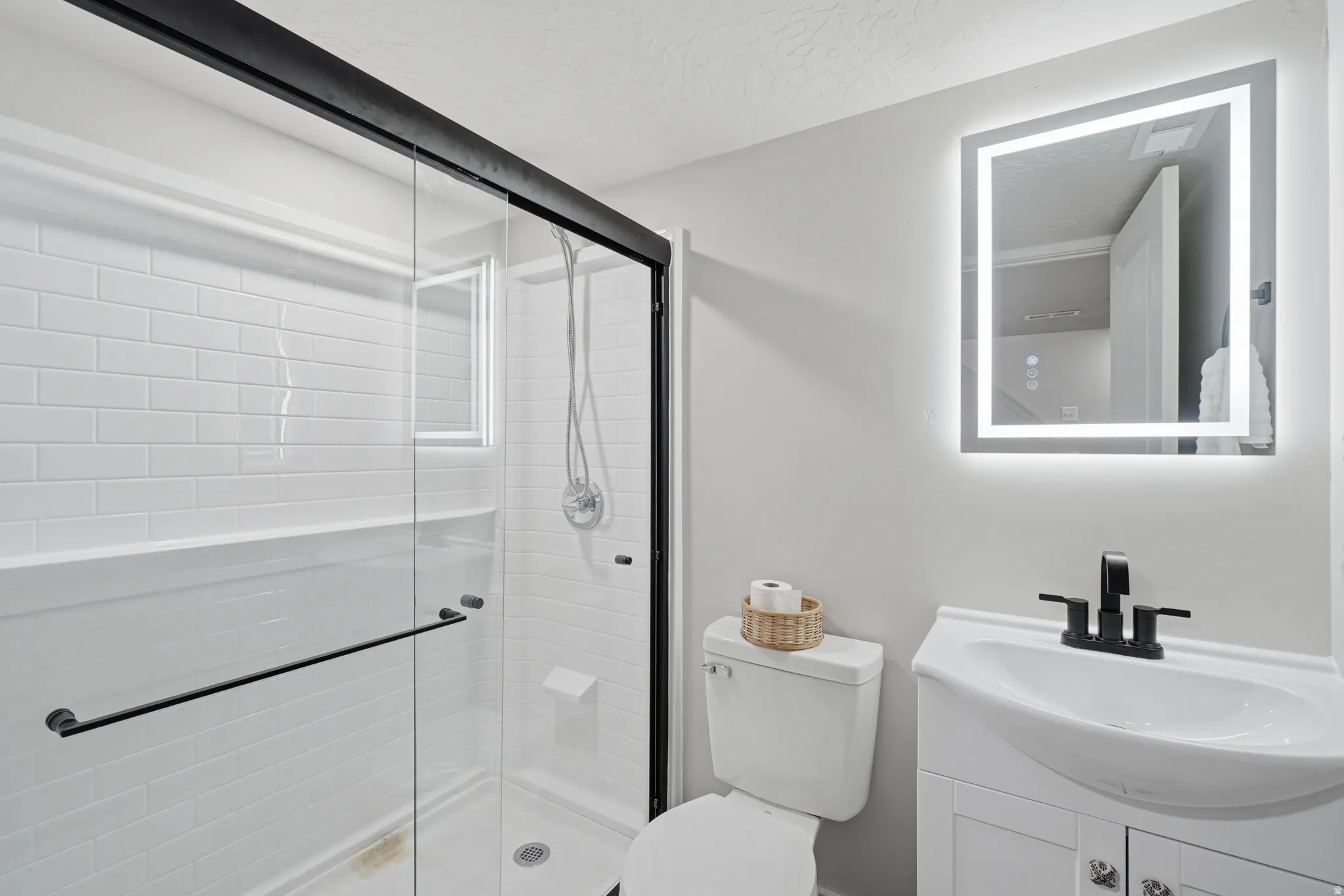 Bathroom featuring an illuminated mirror, white subway tile shower, black framed glass shower enclosure, and a white vanity with black hardware