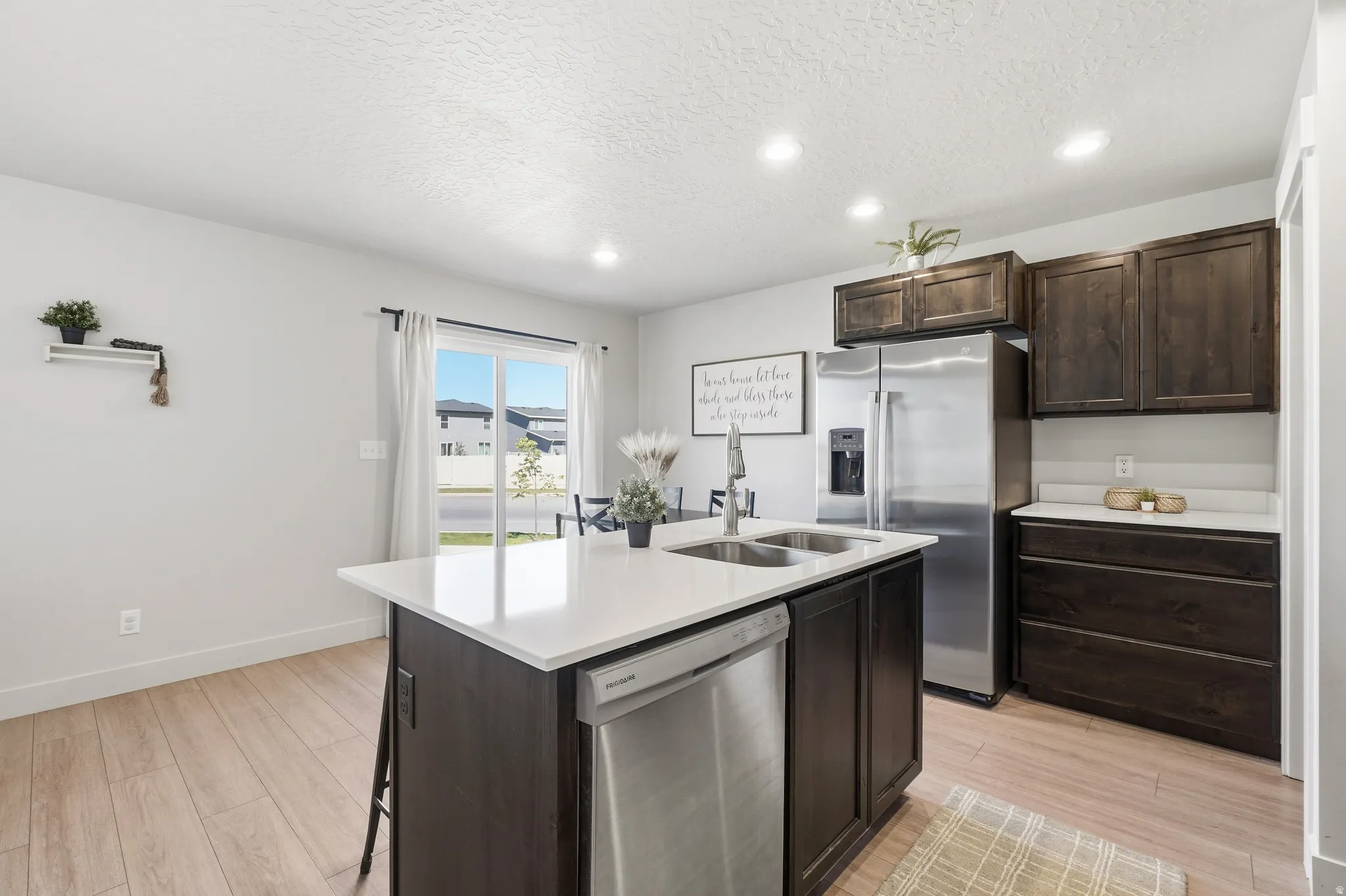 Kitchen featuring wood-finish flooring, dark wood cabinetry, a stainless steel refrigerator, and a kitchen island with a double basin sink and integrated dishwasher