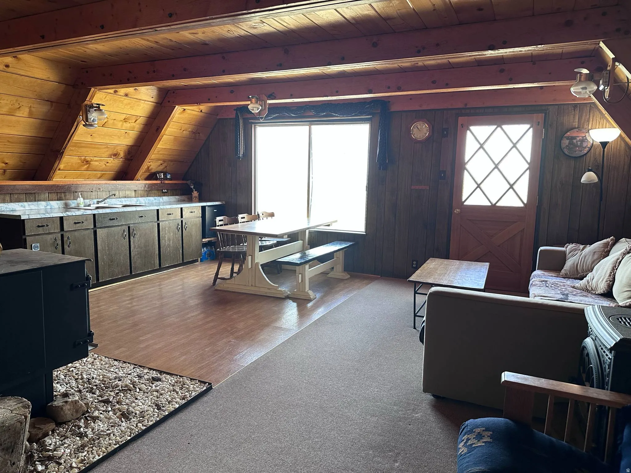 Open-concept living space with wood-paneled walls and ceiling, featuring a wood-burning stove with a stone and wood chip hearth