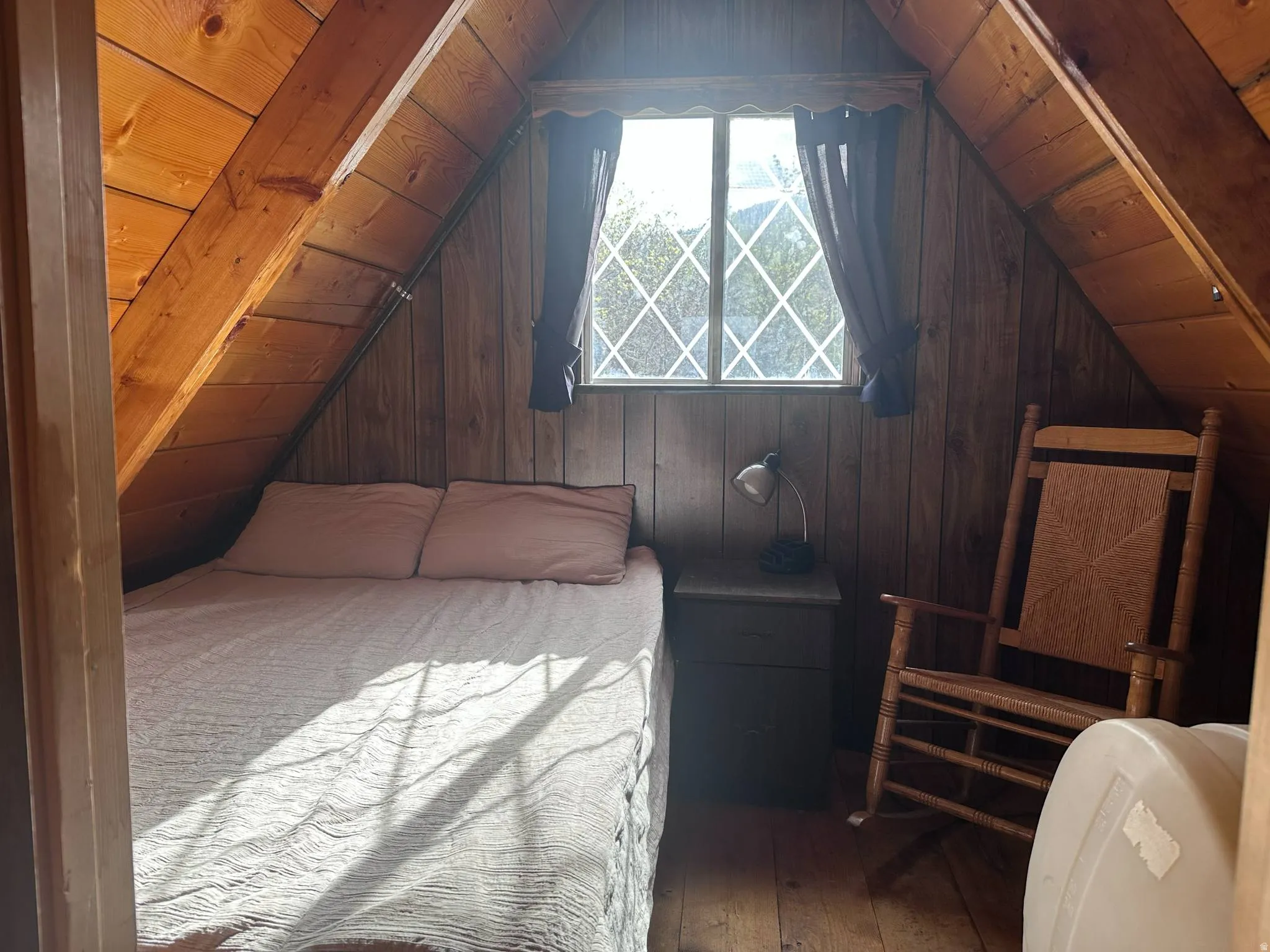 Attic space featuring paneled wood walls, exposed wood beams, diamond-paned window, and wood-finish flooring