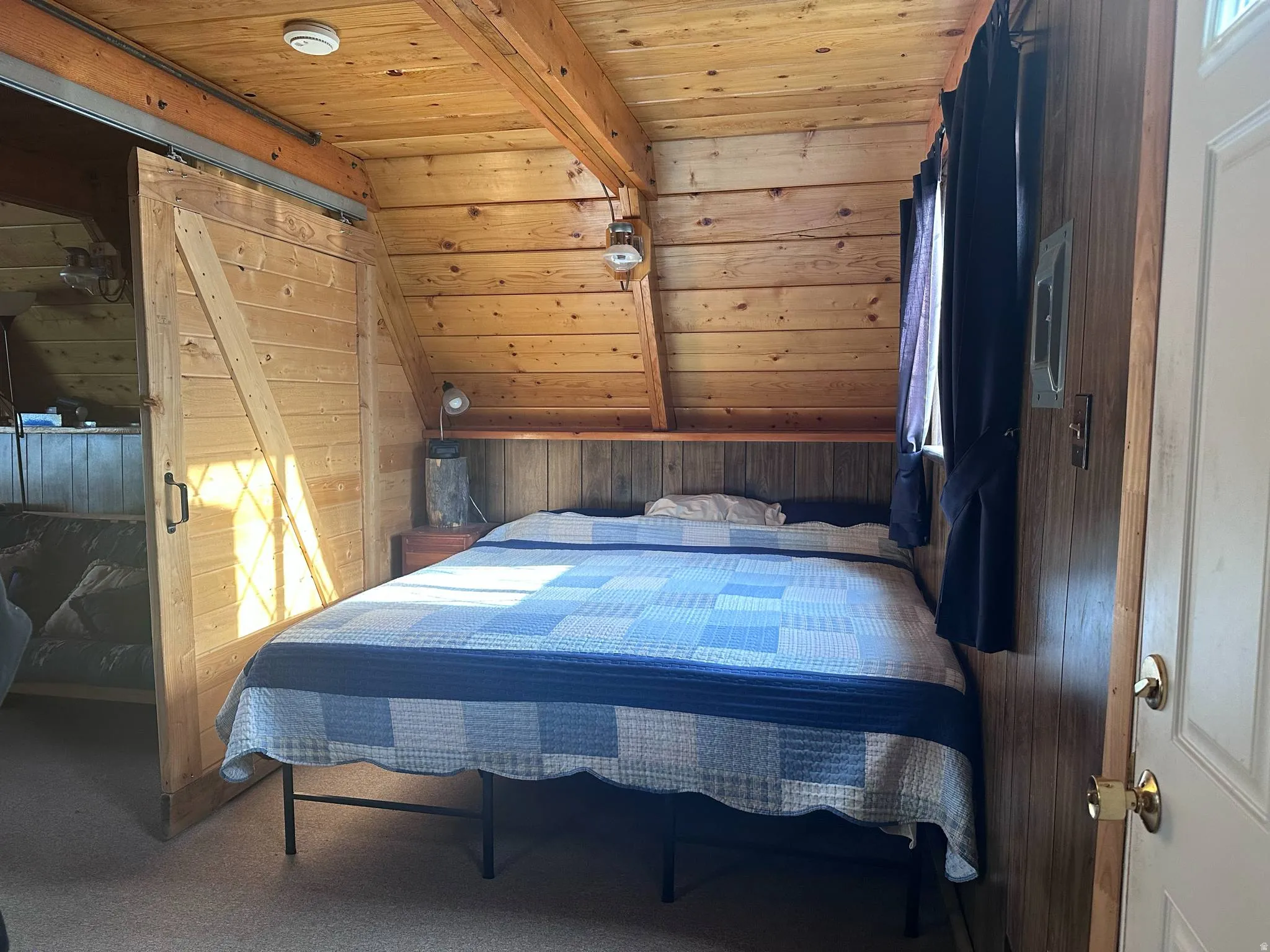 Wood-paneled room featuring a sloped ceiling with exposed beams, a built-in wood headboard, a window with dark drapery, and a wood barn door on a metal track