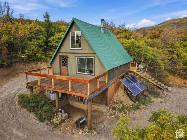 A-frame cabin elevated on wooden pilings, featuring a steep metal roof in forest green, horizontal wood siding, and a wraparound wood-finish deck