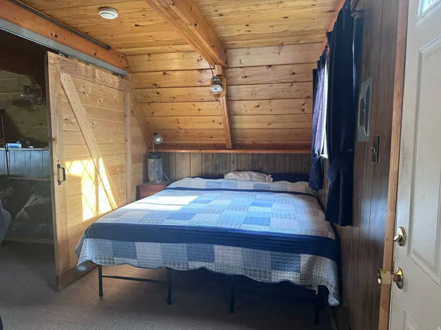 Wood-paneled room featuring a sloped ceiling with exposed beams, a built-in wood headboard, a window with dark drapery, and a wood barn door on a metal track