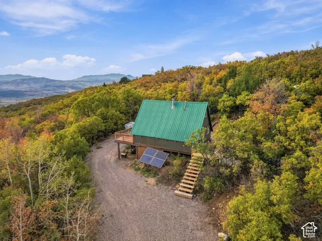 A-frame style residence with a green metal roof, elevated wood deck, exterior solar panels, and wood steps on a natural hillside