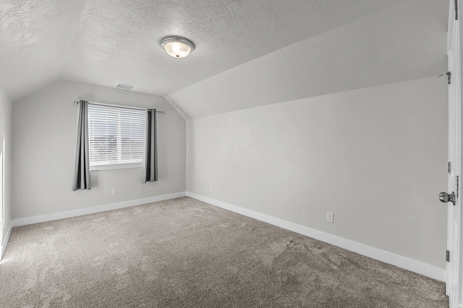Spacious room featuring vaulted ceilings, a large window with blinds, plush gray carpeting, and a flush-mount ceiling light fixture