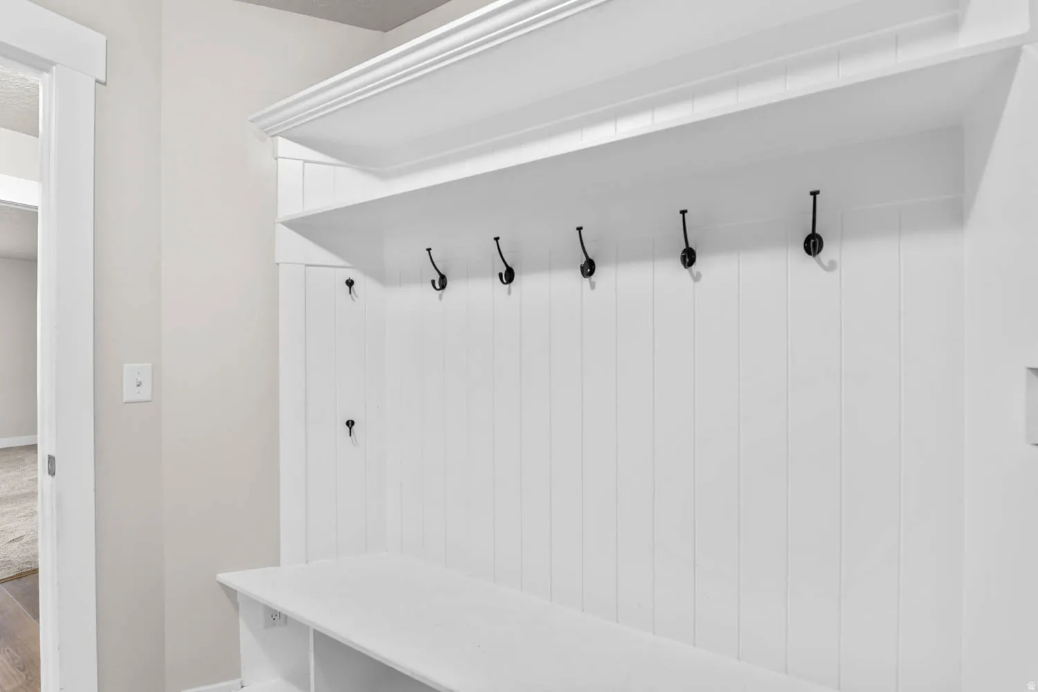 Built-in mudroom bench with cubby storage, vertical white paneling, five black coat hooks, and an overhead shelf with crown molding detail