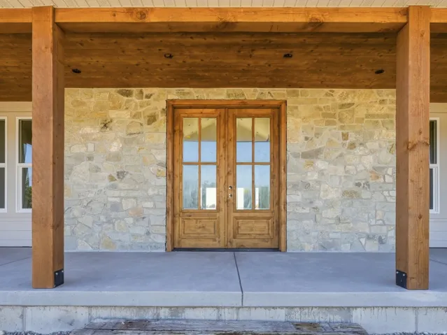Stone facade with a covered concrete porch, featuring exposed wood beams and posts, and a double wood-paneled entry door with glass inserts
