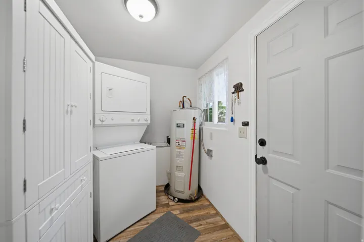 Dedicated utility space featuring wood-finish flooring, white built-in storage cabinetry, a stacked washer and dryer unit, a water heater, and a paneled entry door with black hardware
