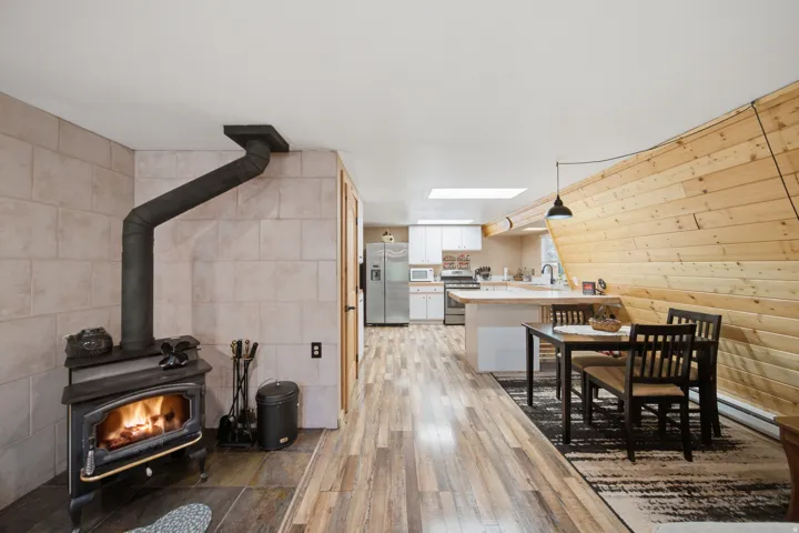Wood-burning stove with a tiled surround, wood-finish flooring, a contemporary kitchen with white cabinetry and stainless steel appliances, a kitchen island with seating, and a sloped wood-paneled wall