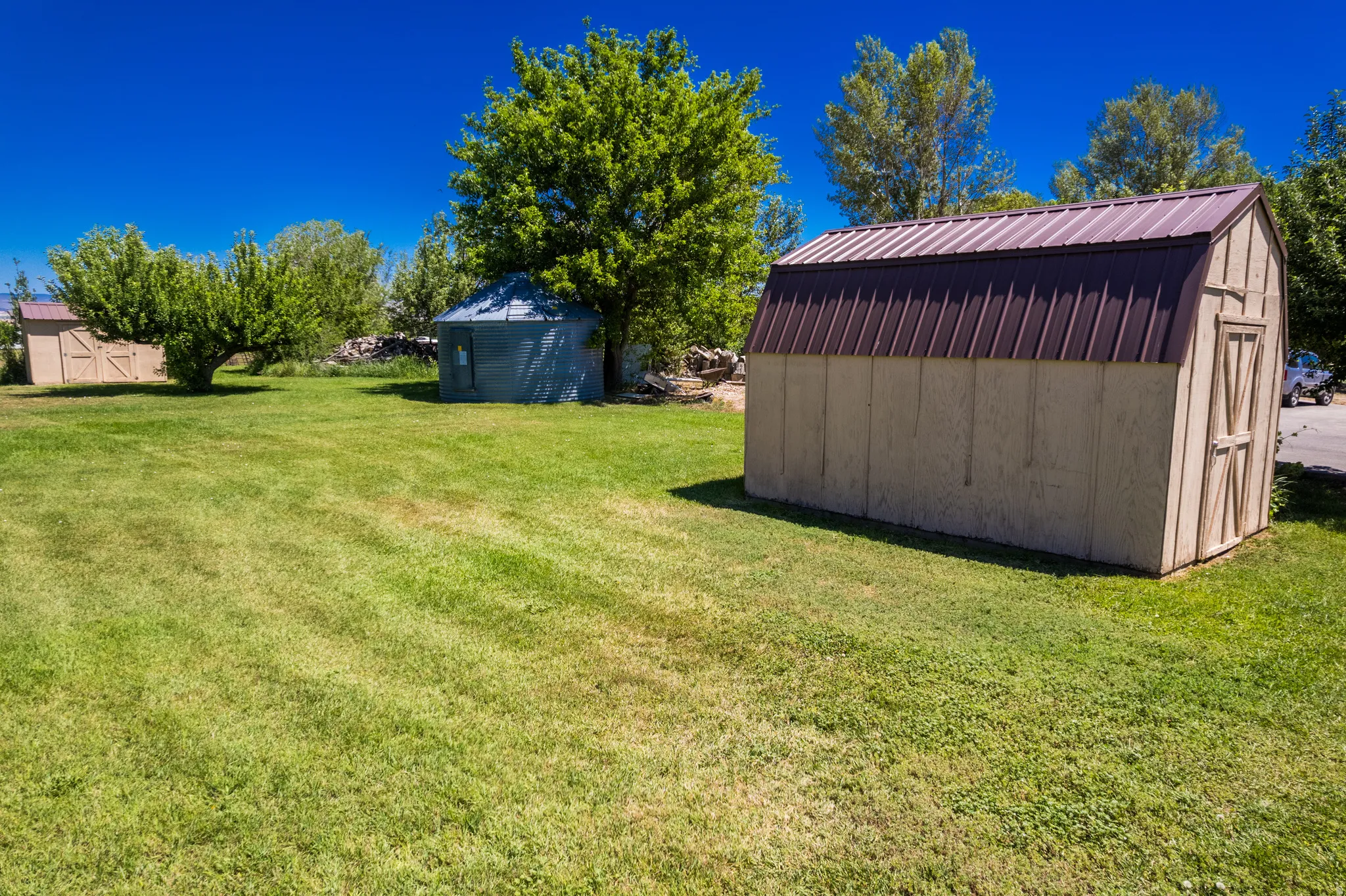 Expansive green lawn with a brown metal-roofed shed, a second storage structure, and mature trees providing natural landscaping
