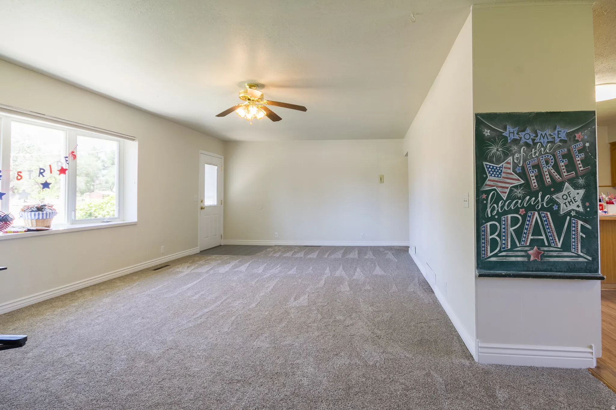Spacious living area featuring neutral carpeting, light wall tones, a ceiling fan with integrated lighting, and a large window providing ample natural light