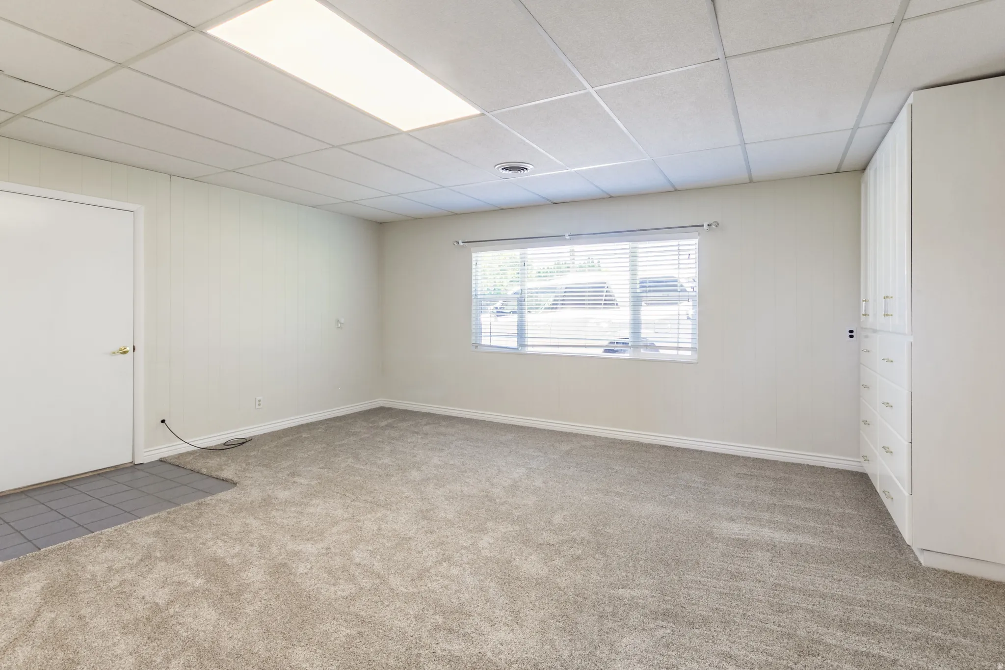 Spacious room featuring neutral carpeting, a large window with horizontal blinds, and extensive built-in cabinetry with multiple drawers and upper storage