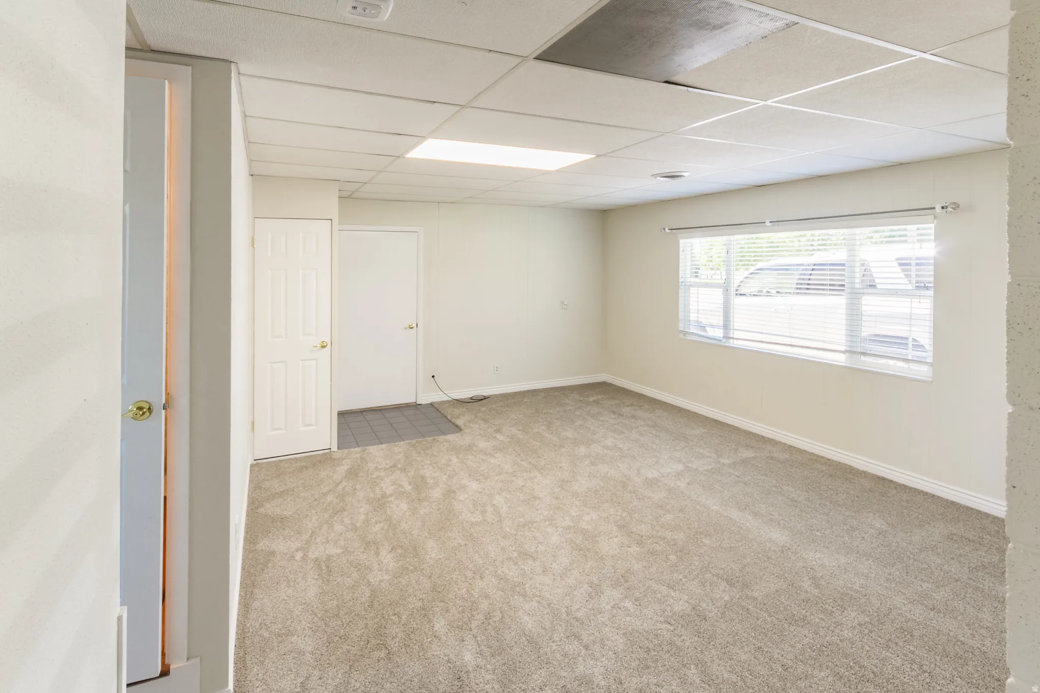 Carpeted room featuring a large window with blinds, drop ceiling, and multiple doors