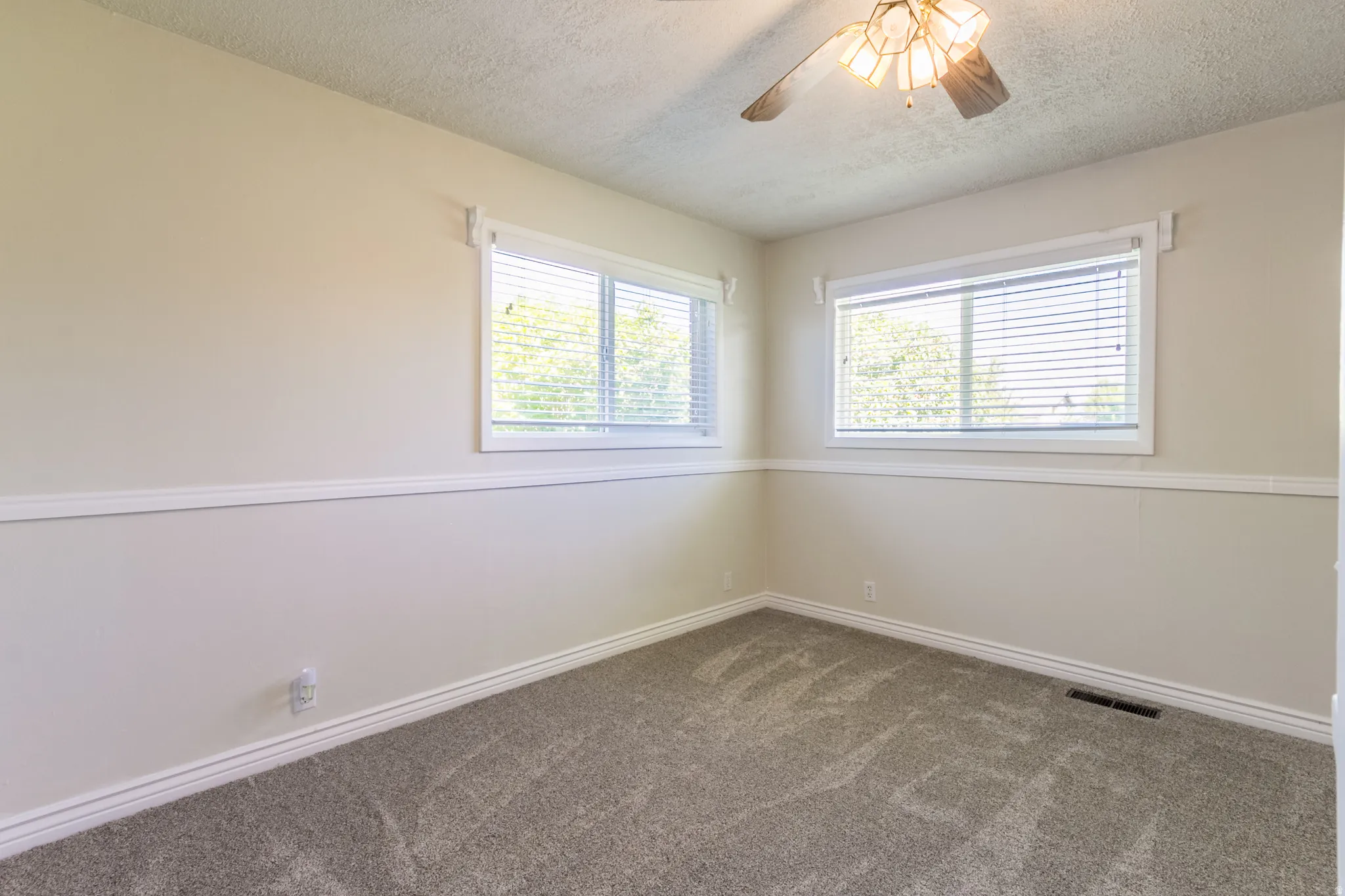 Neutral-toned room featuring a ceiling fan with integrated lighting, two windows with blinds, white trim, and patterned carpeting