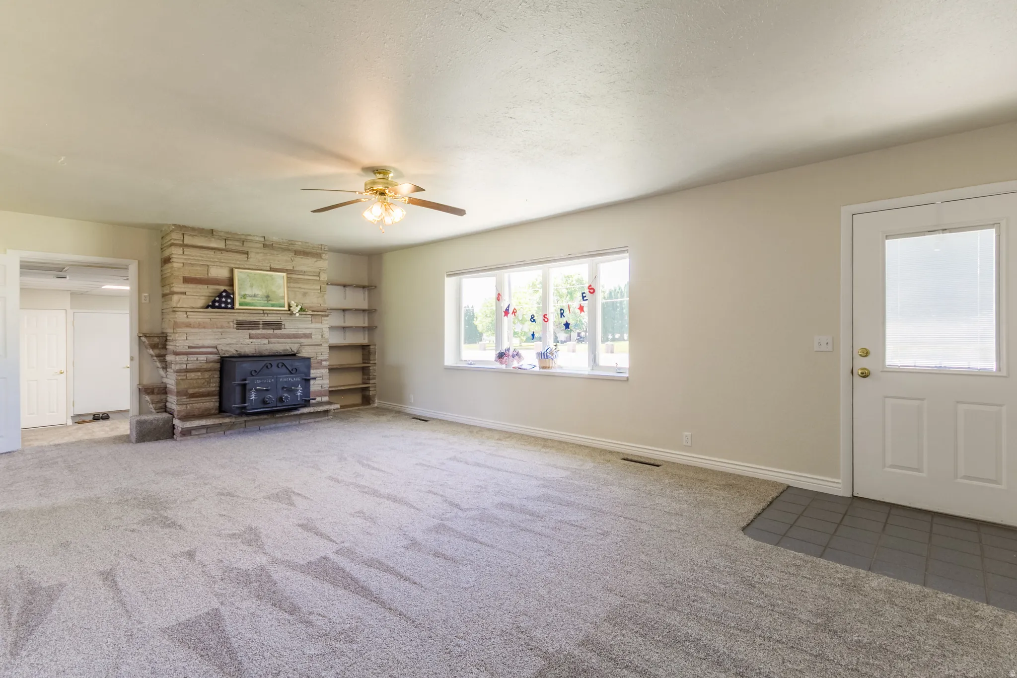 Spacious living area featuring a stone fireplace with a wood-burning insert, built-in shelving, a ceiling fan, a large window, and light-colored carpeting