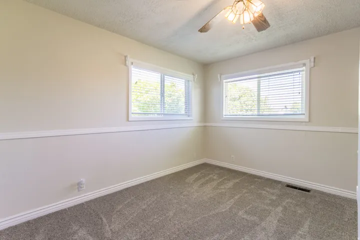 Neutral-toned room featuring a ceiling fan with integrated lighting, two windows with blinds, white trim, and patterned carpeting